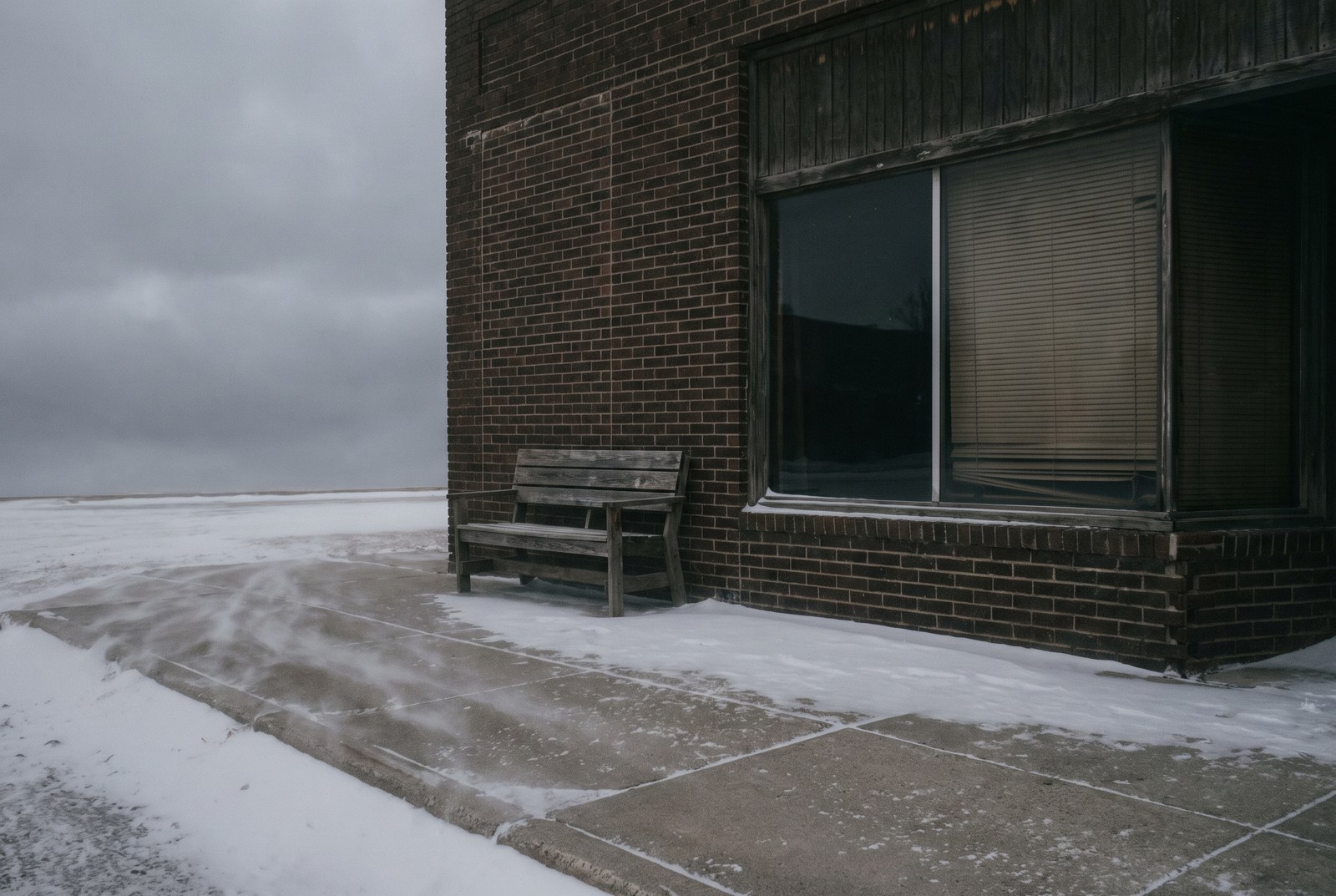 A small-town brick storefront with drawn blinds and a bench outside while snow blows across the sidewalk.