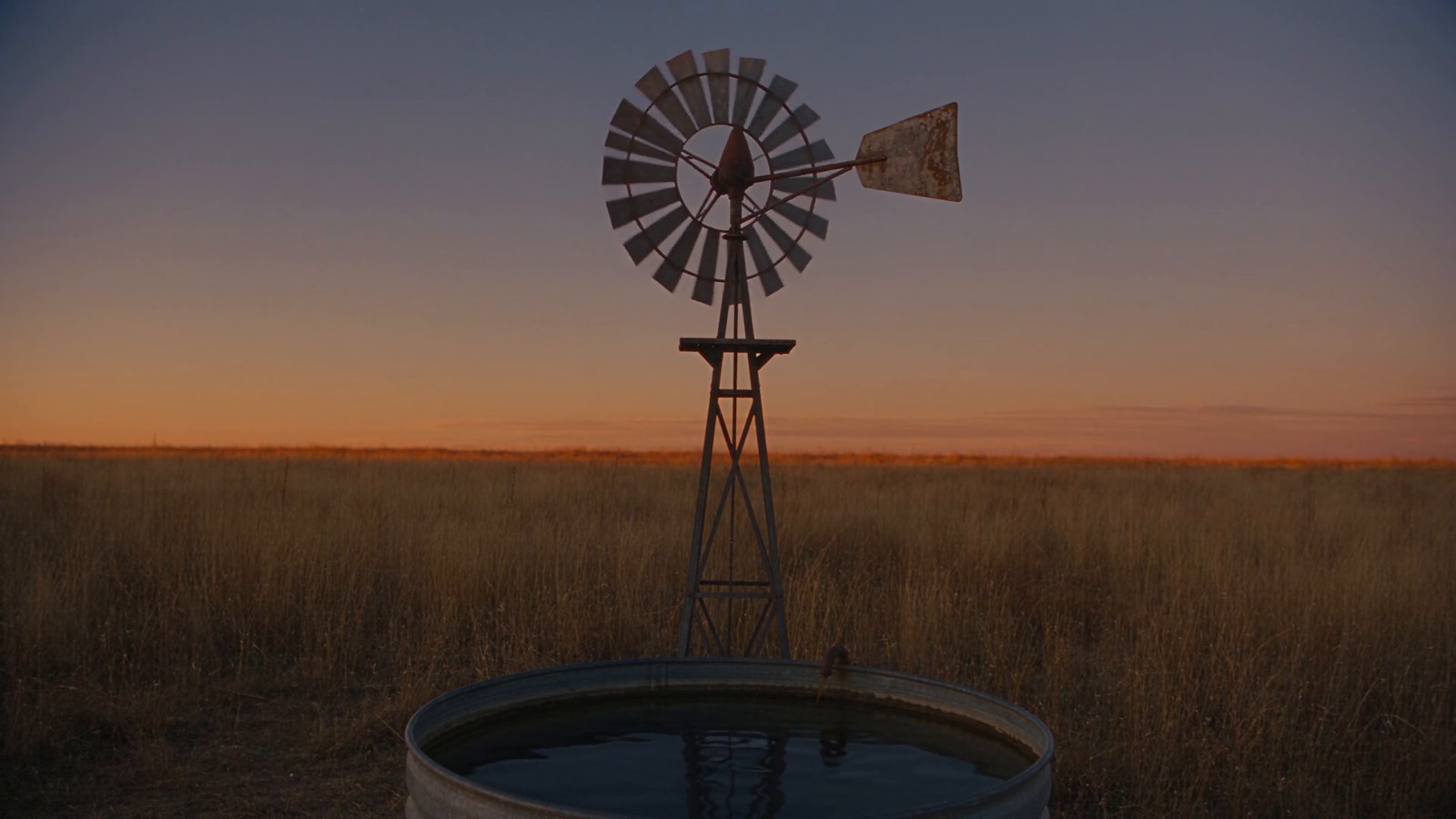 A windmill water pump turns slowly over a stock tank on the prairie at sunset.