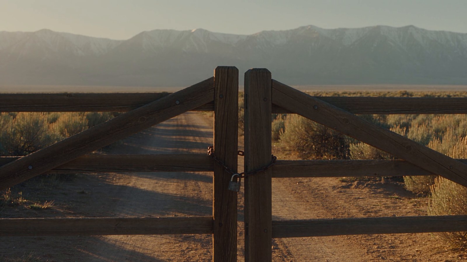 A chained wooden ranch gate across a dirt road that runs toward distant snowy mountains.